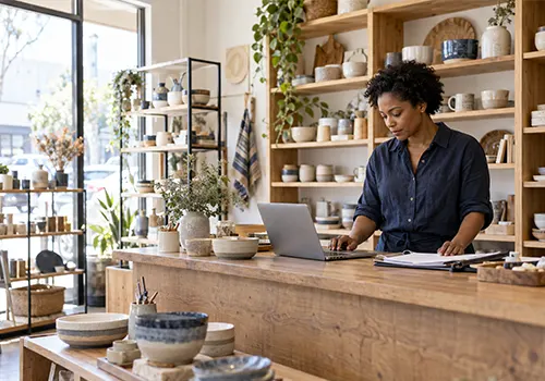 Small business owner reviewing paperwork and using a laptop behind the counter of a ceramics shop.